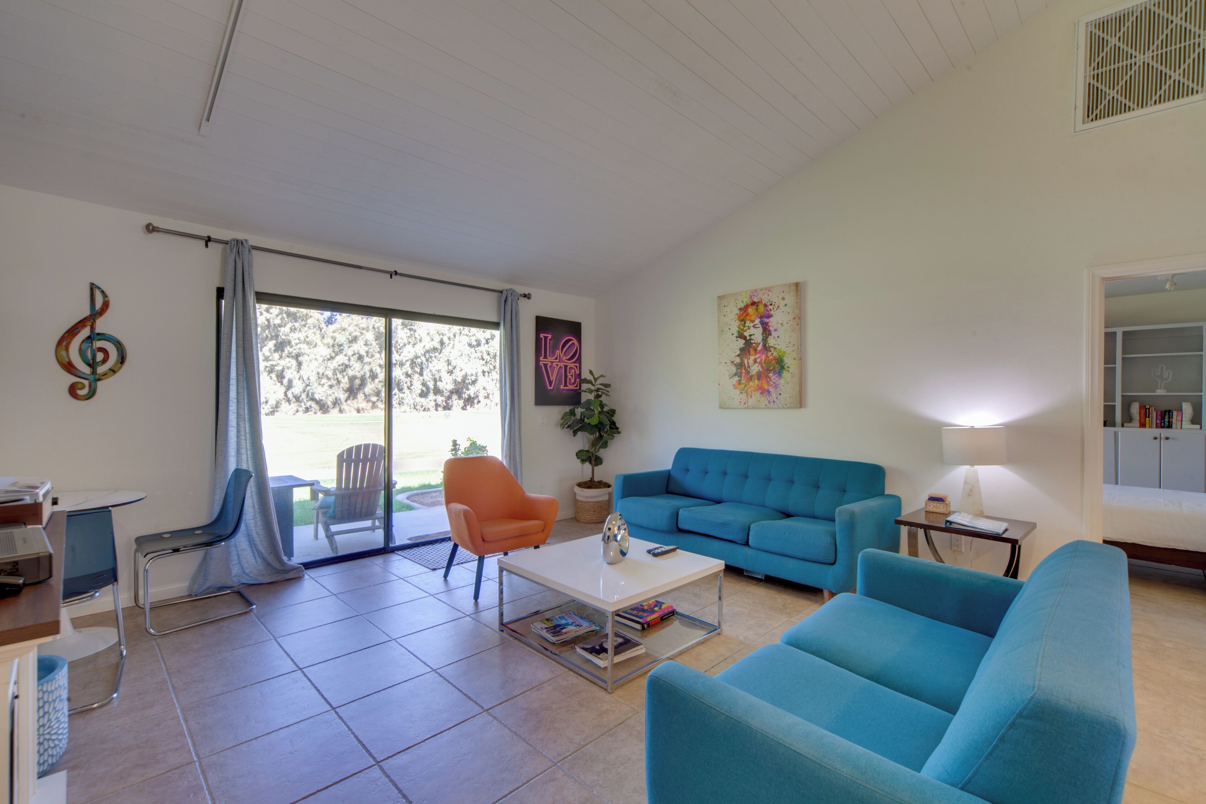 A living room with a blue sofa, a coffee table, and a chair. There is a window with curtains, a plant, and a painting on the wall.