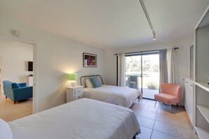 A bedroom with two beds, a chair, a bedside table with a lamp, and a window with curtains.