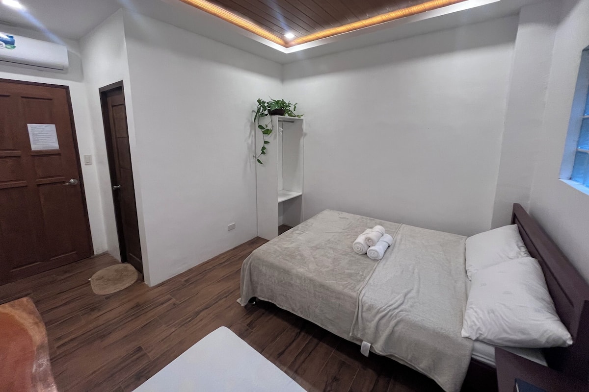 Un dormitorio con una cama, suelo de madera, pared blanca, techo con iluminación empotrada, una puerta y una ventana.