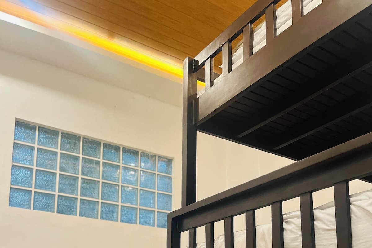 Una cama litera con estructura de madera y ropa de cama blanca, una ventana con cristales azulados y suelo de madera.