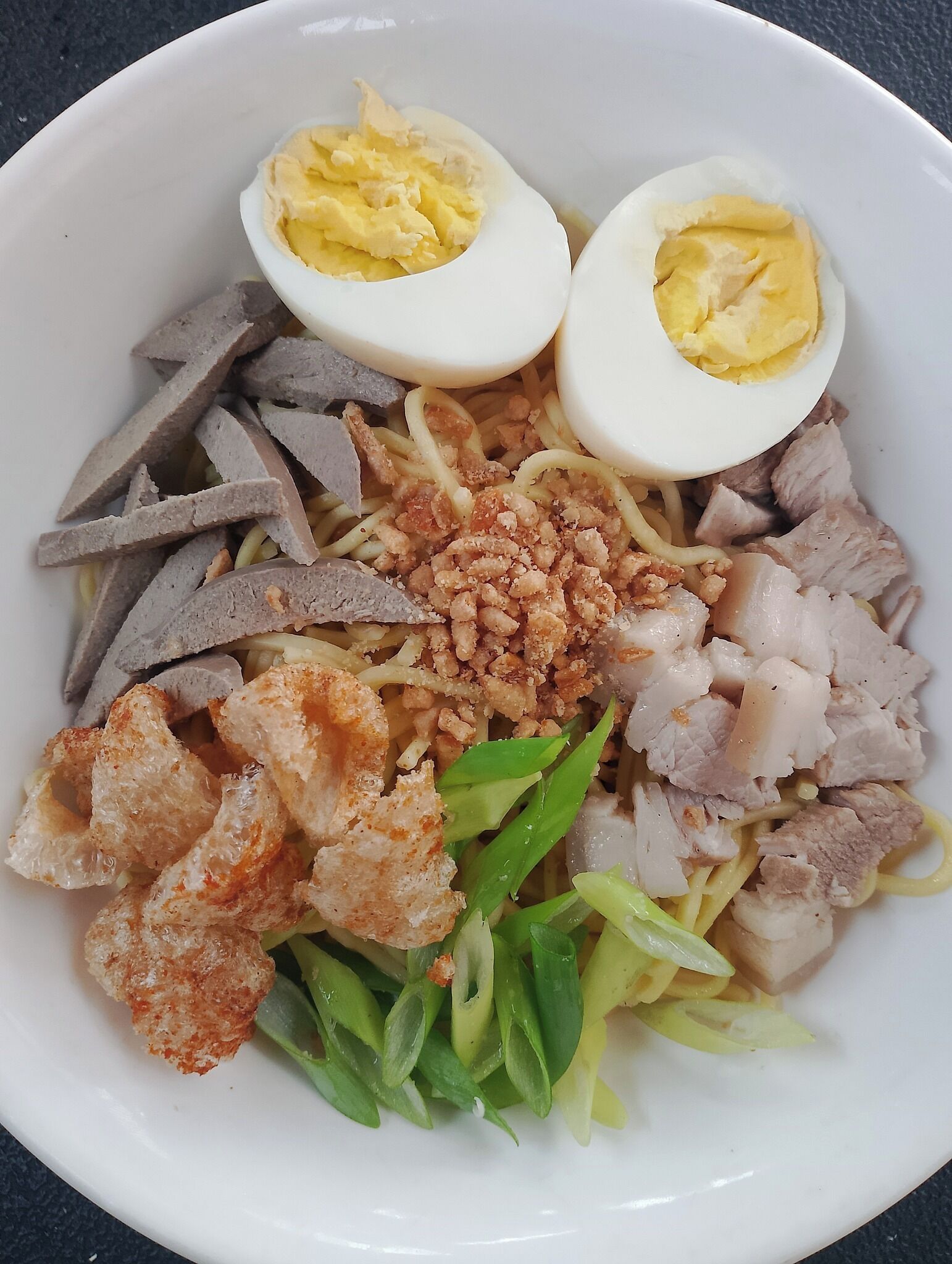 A bowl of noodle dish with sliced meat, fried items, and boiled eggs.