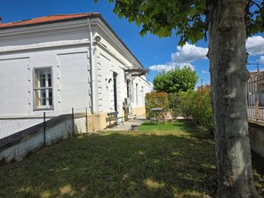 A white building with a red-tiled roof, a small garden with a bench, and a tree.