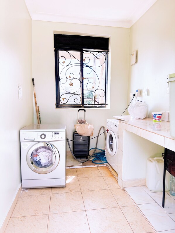A laundry room with a washing machine, dryer, and a suitcase. - Rodland Furnished Apartments (Kampala)