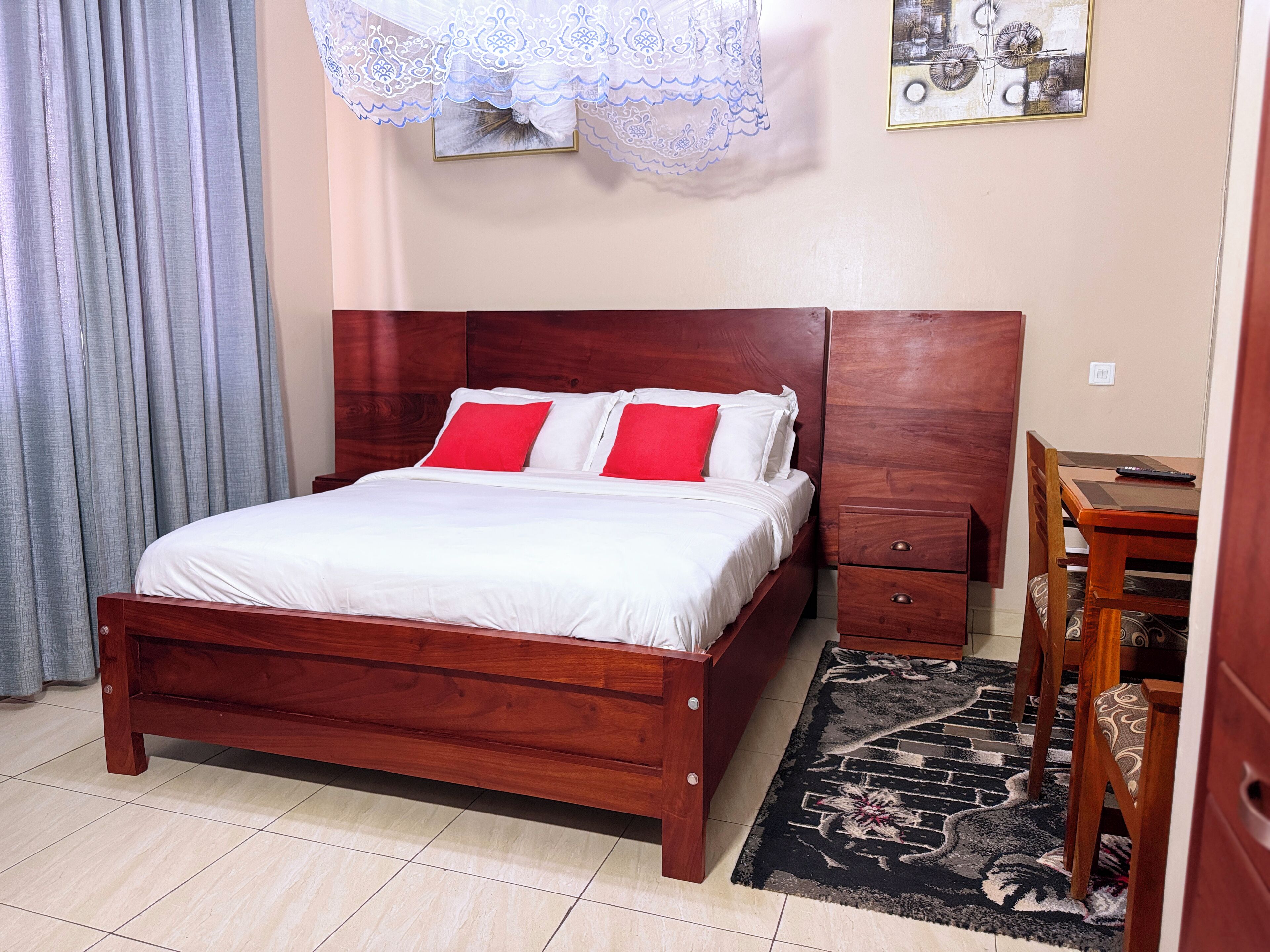 A neatly made bed with white and red bedding, a wooden headboard, a small bedside table, and a chair with a patterned cushion.