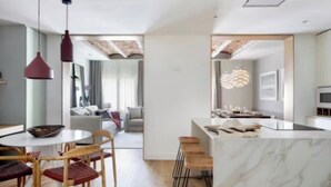 Una cocina moderna con encimera de mármol, taburetes de madera, iluminación colgante y vistas al salón.