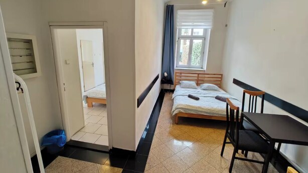 Habitación pequeña con cama, una mesita y una silla. Hay una puerta que da a otra habitación.