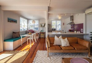 A modern living space with a dining area, a kitchen, and a seating area with a sofa and cushions. - The Cottage – Oneroa’s Best Viewpoint (Waiheke Island)