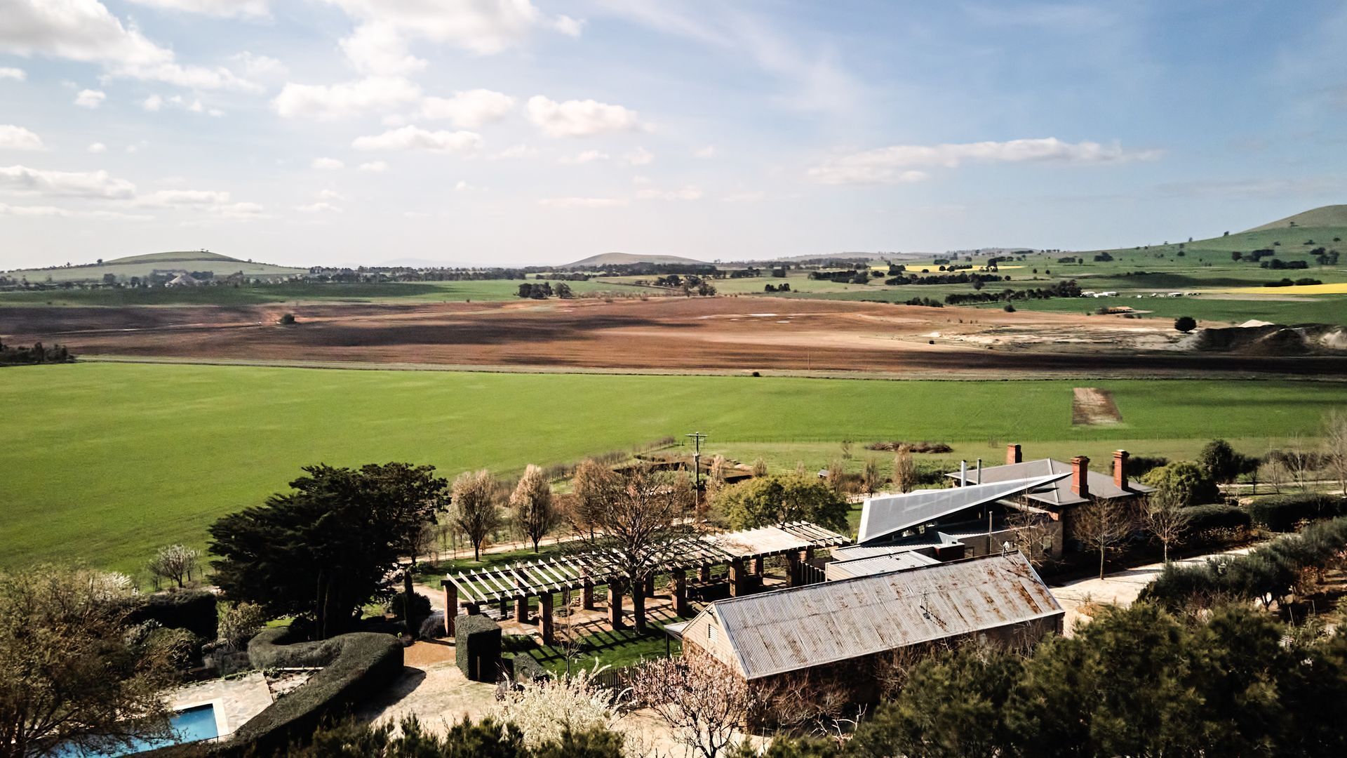 Un paysage rural avec un grand champ verdoyant, une pergola en bois, un bâtiment doté d’un toit en métal et une piscine.