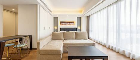A modern living room with a sectional sofa, a dining table with chairs, and a large window with sheer curtains.