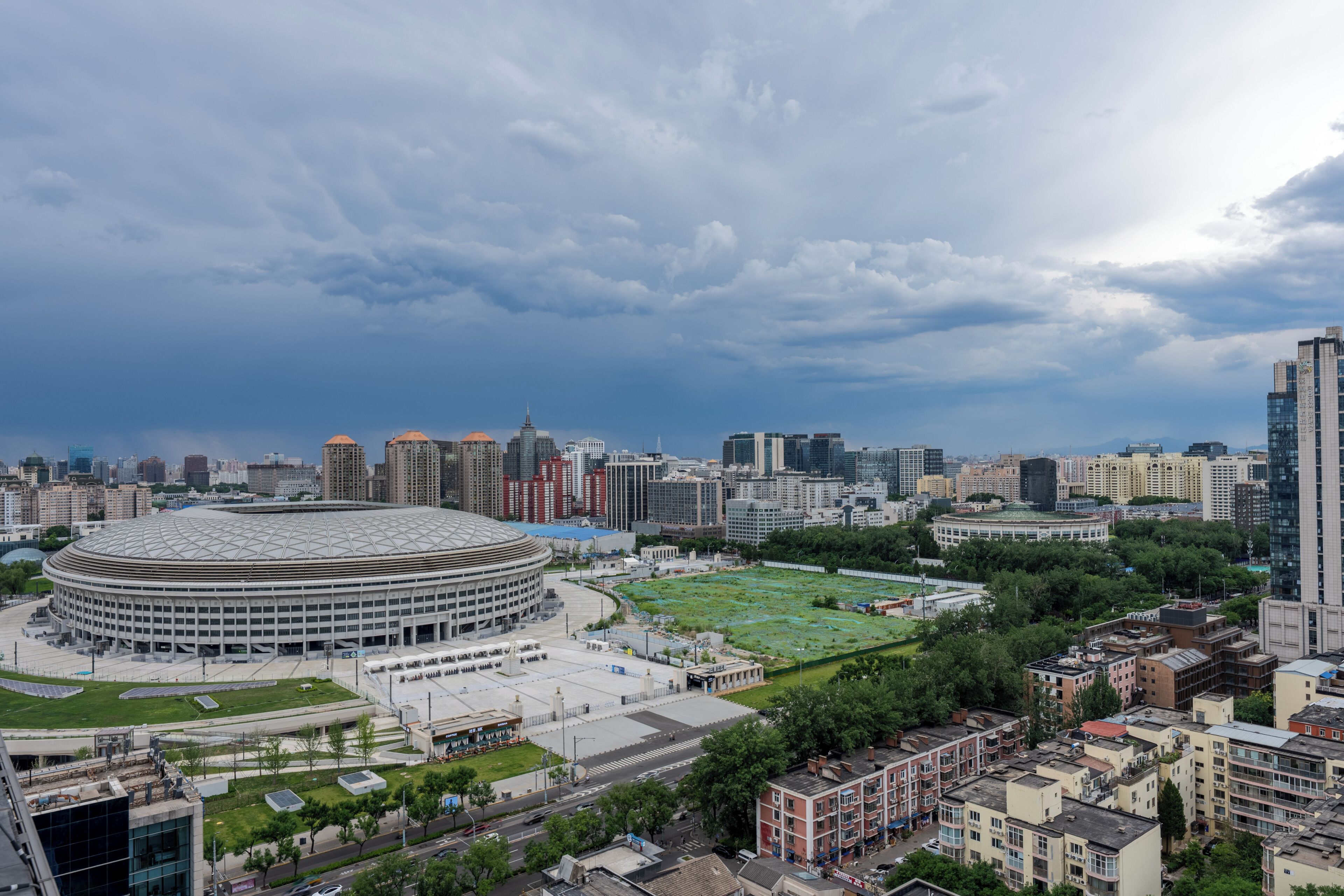 A cityscape with a large circular building, surrounded by various other buildings and a green park area.