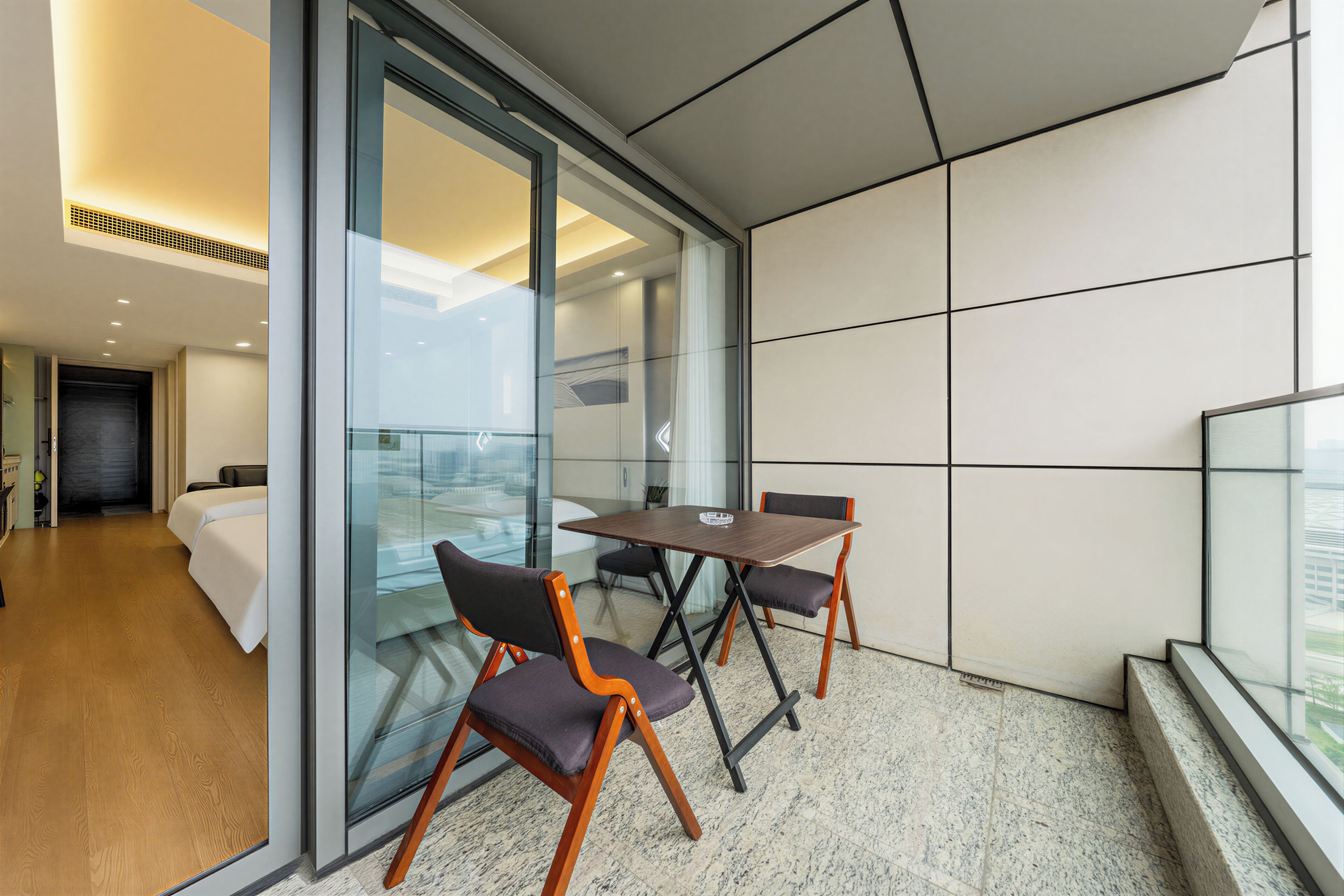 Ein modernes Hotelzimmer mit Glaswand, Holzboden, einem Bett und einem Essbereich mit Stühlen und Tisch.