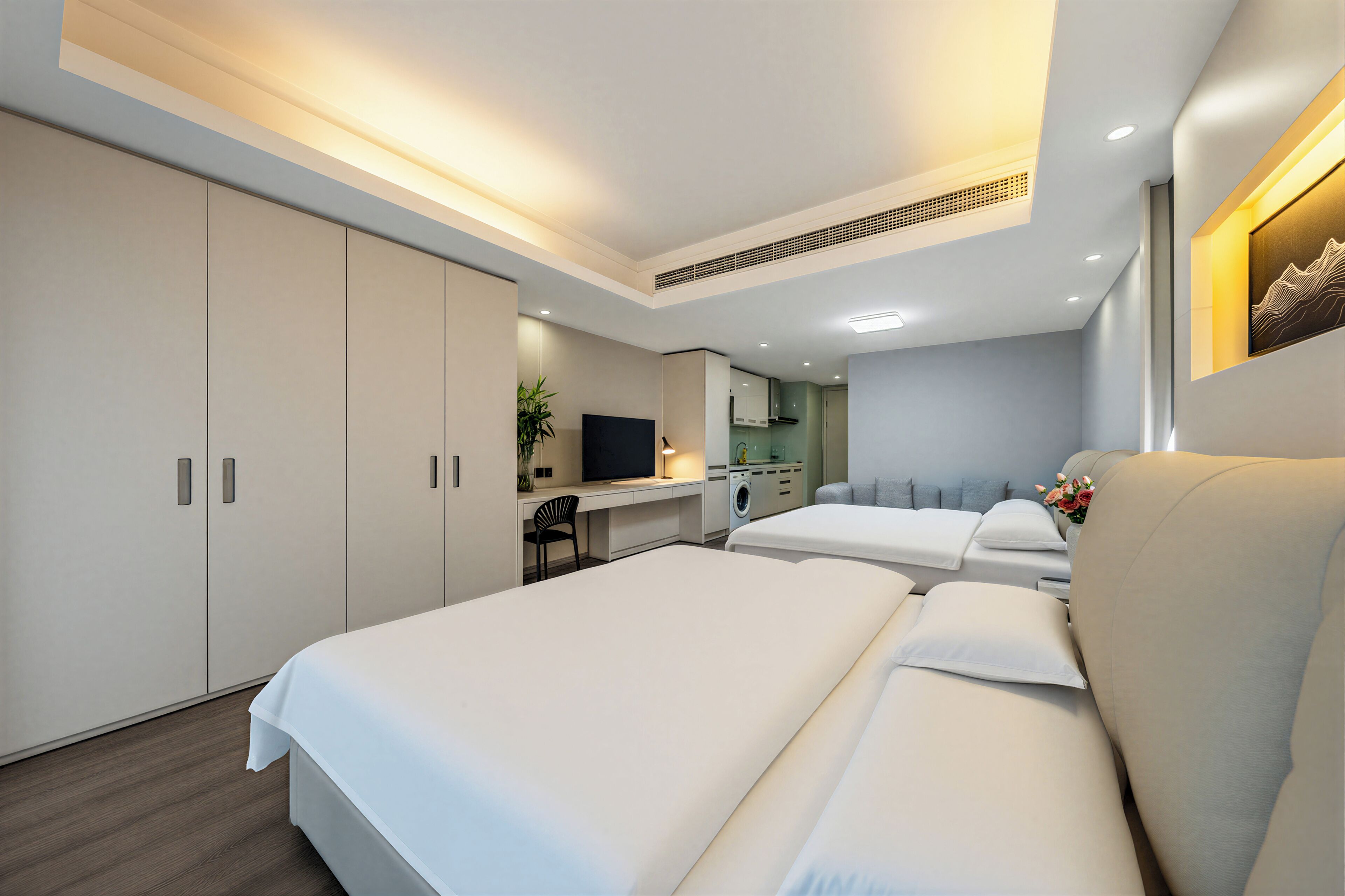 A modern hotel room with a large bed, a desk with a chair, a washing machine, and a small kitchenette.