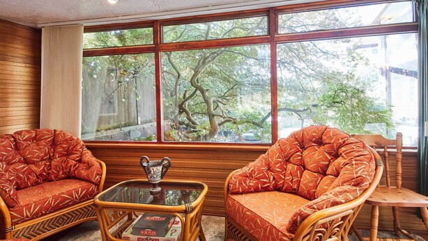 A room with two wicker armchairs, a glass coffee table with a Monopoly game, and a view of trees through large windows. - Host & Stay | Windles (Wotton-under-Edge)
