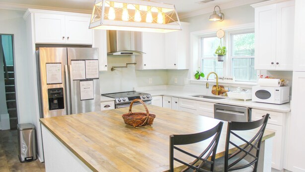 Una cocina moderna con armarios blancos, electrodomésticos de acero inoxidable, una isla de madera con sillas y una lámpara colgante.