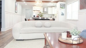 Una sala de estar moderna con un sofá blanco grande, una mesa de centro de madera y una cocina al fondo.