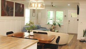 Un área de comedor con una mesa de madera, sillas y un jarrón con plantas. Hay un sofá y una ventana con vistas a la vegetación.