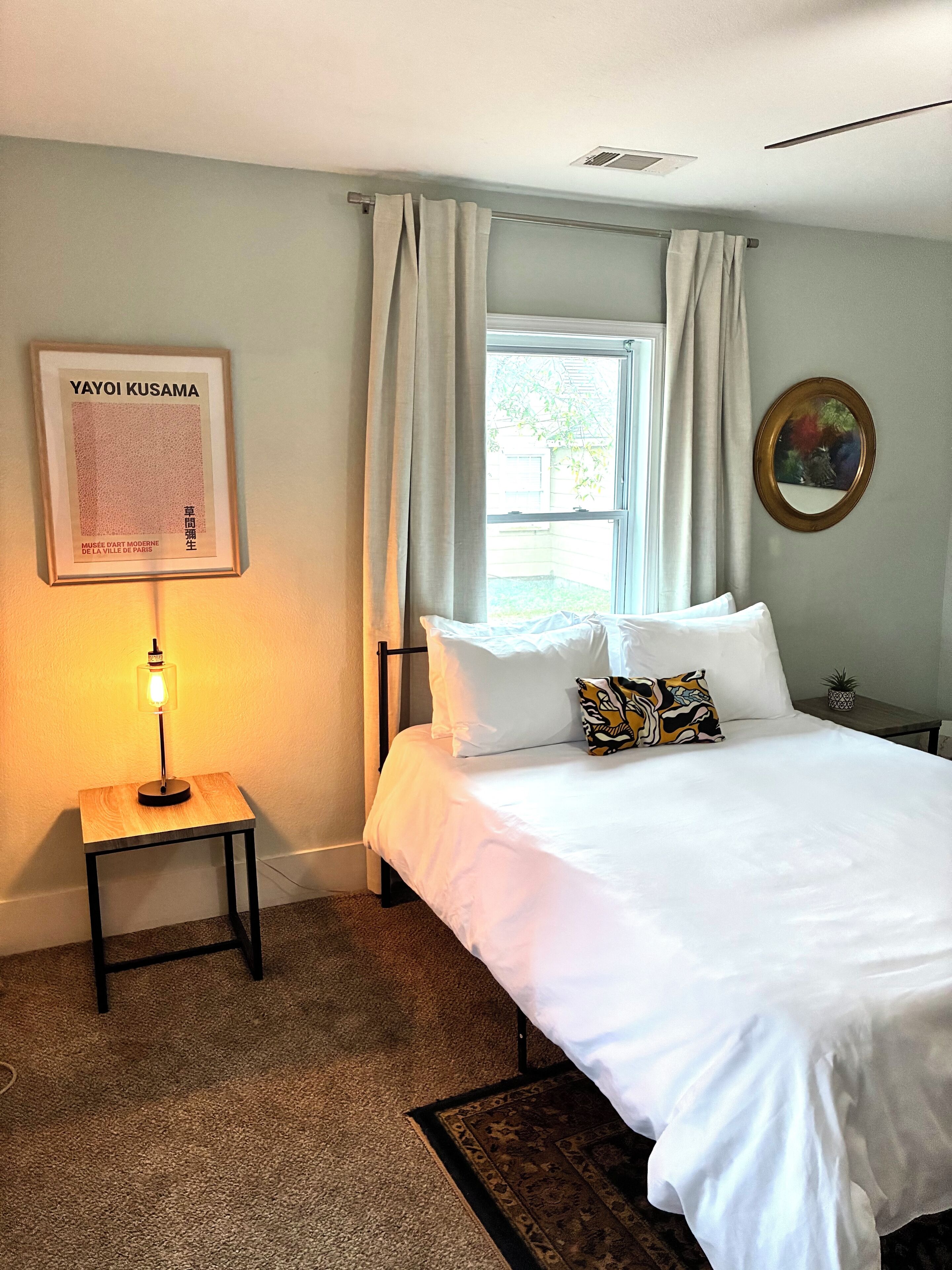 A hotel room with a bed, a nightstand with a lamp, a framed artwork, a window with curtains, and a small potted plant.