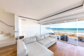 A modern living room with a large sectional sofa, wooden flooring, and a view of the beach through floor-to-ceiling windows.