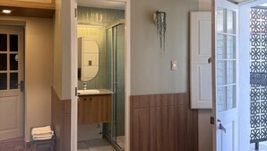 Une chambre avec un lit, un petit tabouret, une salle de bain avec un miroir et une porte donnant sur l’extérieur.