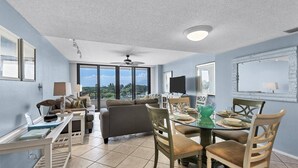 A spacious living area with a dining table set for four, a sofa, a television, and a large window offering a view of the outdoors.