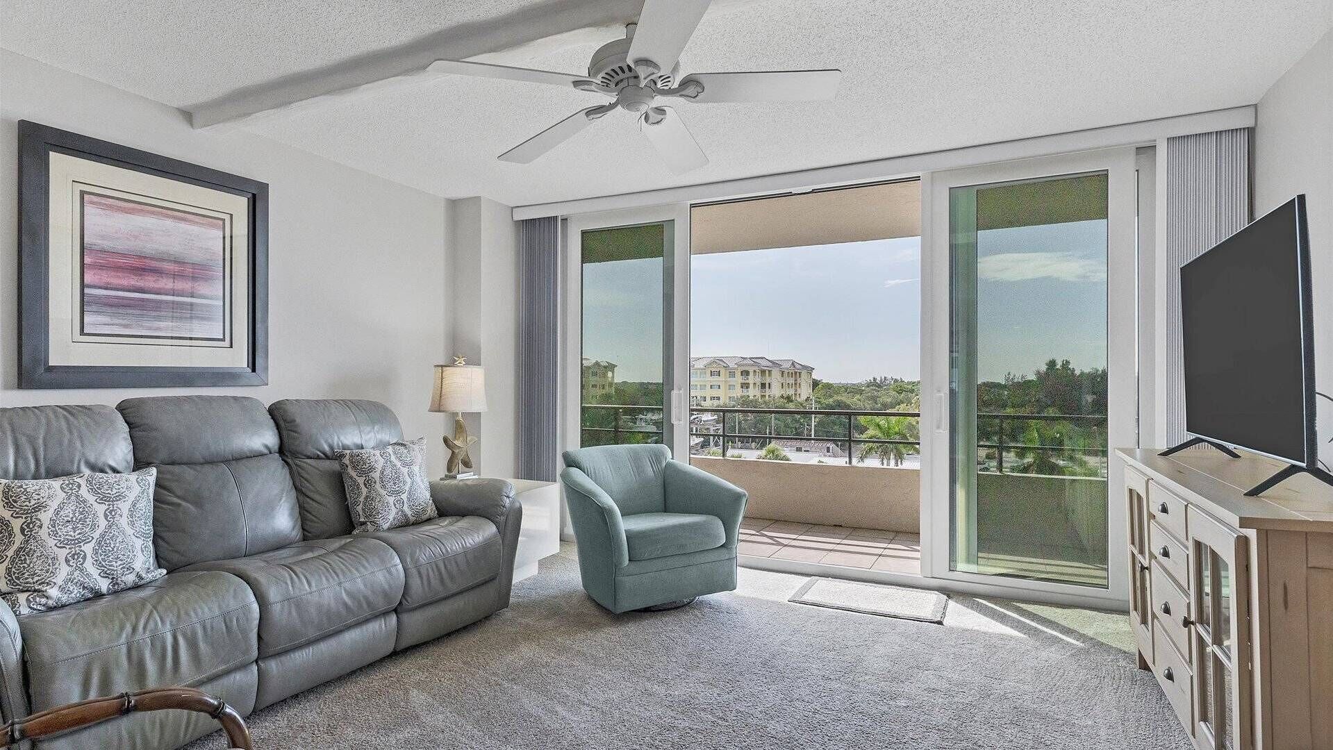 A living room with a grey leather sofa, a blue armchair, a ceiling fan, and a large window offering a view of buildings and greenery.
