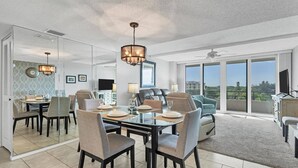 A modern dining area with a glass table, chairs, and a chandelier. There is a living area with a sofa and a TV. The room has large windows and a ceiling fan.