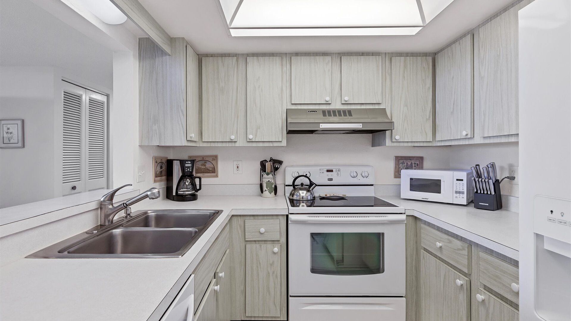 Una cocina moderna con armarios blancos, un lavamanos de acero inoxidable, una cafetera, un hervidor, un microondas y un horno.