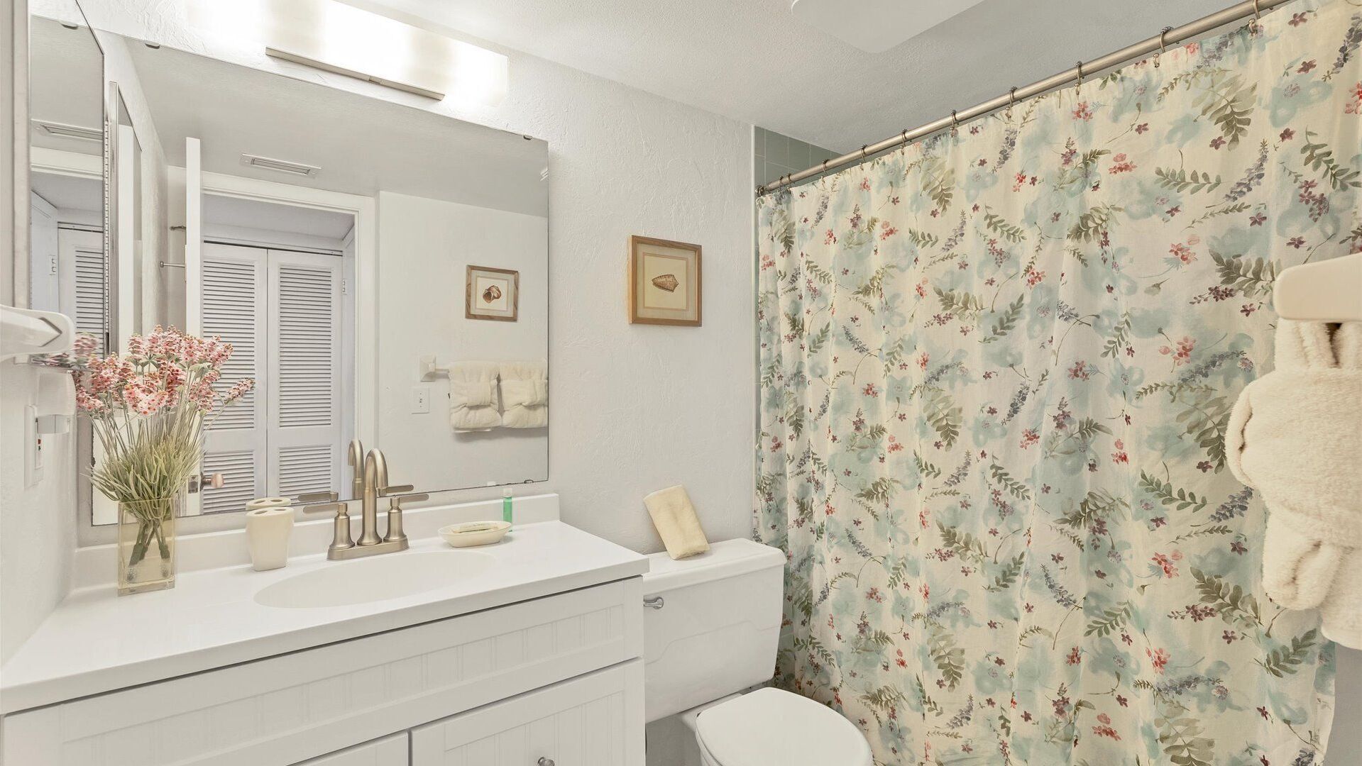 A bathroom with a white vanity, a large mirror, a shower curtain with a floral pattern, and a toilet.