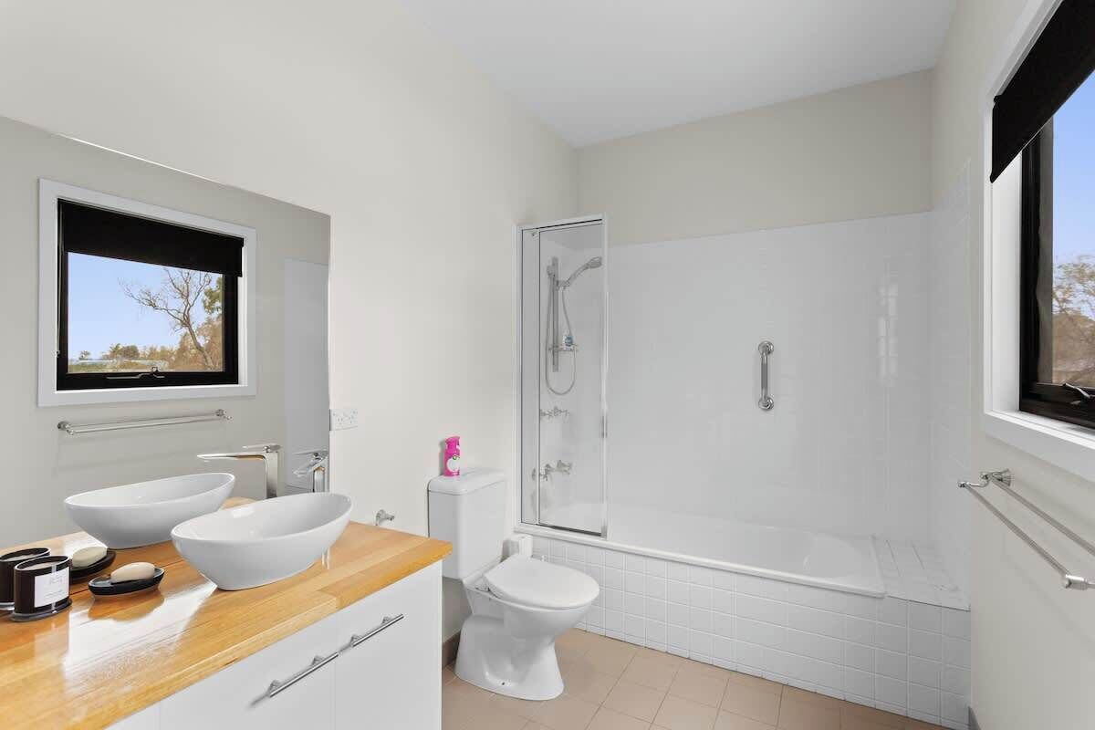 A modern bathroom with a wooden vanity, two white sinks, a toilet, and a glass-enclosed shower.