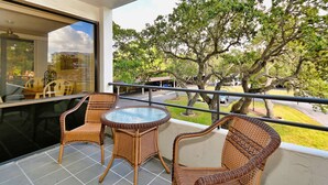 A balcony with wicker furniture, a glass table, and a view of trees and a road.