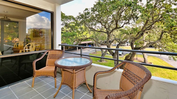 A balcony with wicker furniture, a glass table, and a view of trees and a road.