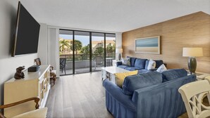 A modern living room with a blue sofa, a flat-screen TV, and a balcony with a view of palm trees and buildings. - Anchorage Resort 2 Bedroom Condo 2nd Floor, Community Pool 0883 (Siesta Key)