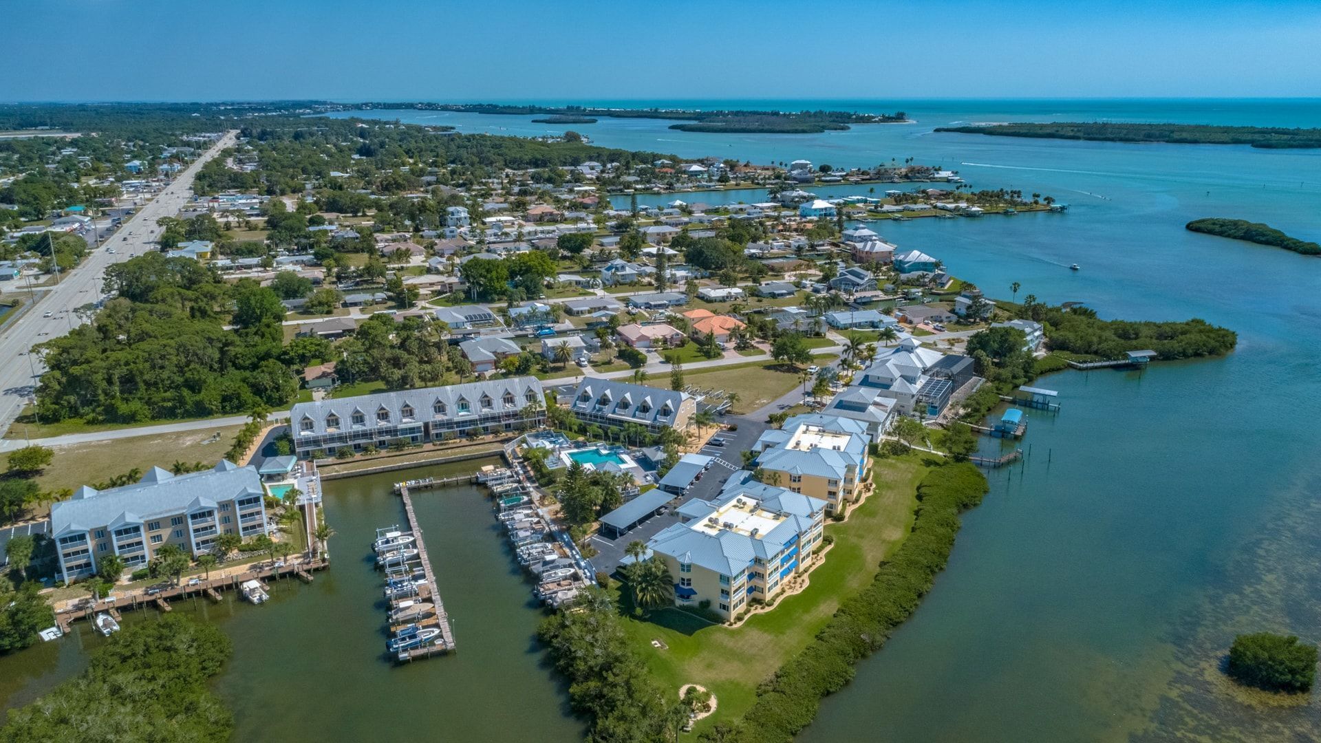 An aerial view of a coastal residential area with houses, a marina, and a large building with a swimming pool.