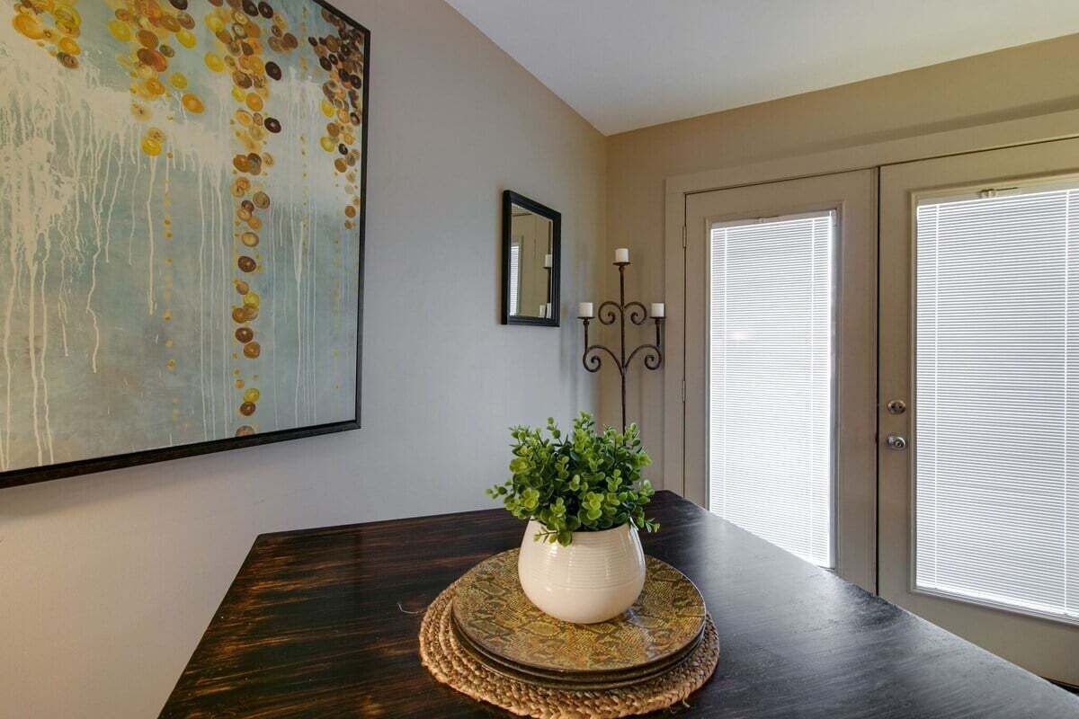 Un coin repas avec une table en bois foncé, un vase blanc contenant des plantes vertes, un grand tableau abstrait, un miroir et une porte avec des stores.