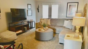 A living room with a television, sofa, ottoman, and a small side table with a lamp. - Loft #304 (540 N 2nd St) (Clarksville)
