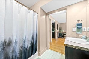 A bathroom with a shower curtain depicting a forest scene, a sink with a granite countertop, and a mirror with a towel hanging on the wall. - Loft #105 (540 N 2nd St) (Clarksville)