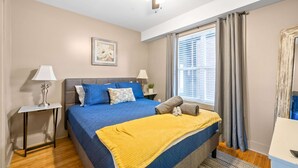 Ein Schlafzimmer mit einem Bett, einem Deckenventilator, einem Fenster mit Vorhängen, einem Nachttisch mit Lampe und einem Wandbild.