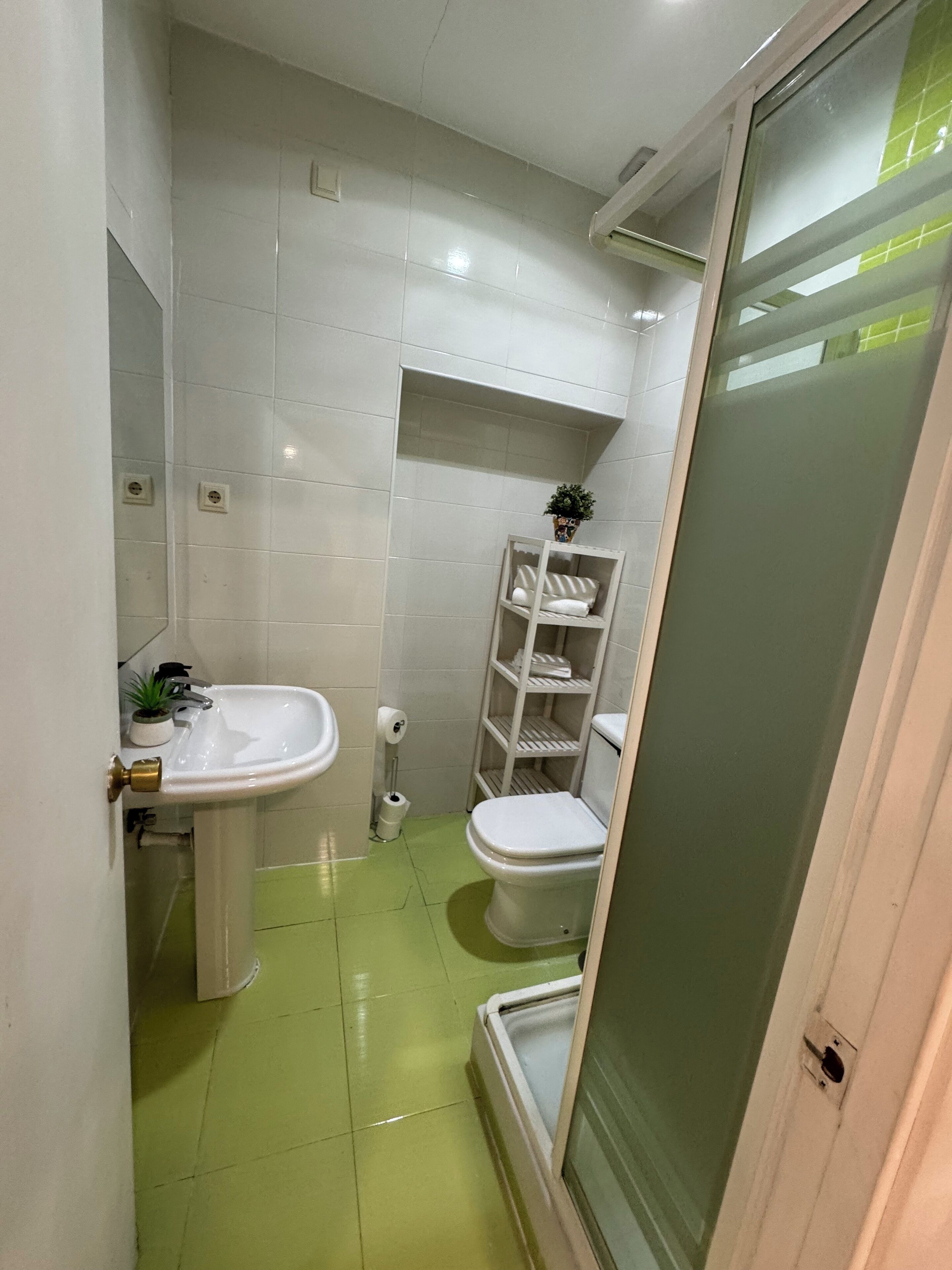 Un baño con azulejos blancos, un lavabo, un inodoro y un estante con artículos de aseo.