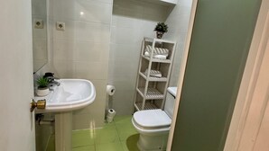 Une salle de bain avec des carreaux blancs, un lavabo, des toilettes et une étagère contenant des articles de toilette.