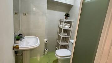 Un baño con azulejos blancos, un lavabo, un inodoro y un estante con artículos de aseo.