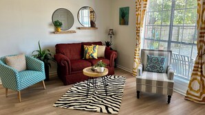 A living room with a red sofa, a blue patterned armchair, a zebra-patterned rug, and a striped armchair by a large window with curtains.