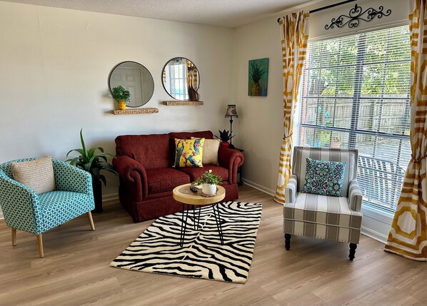 A living room with a red sofa, a blue patterned armchair, a zebra-patterned rug, and a striped armchair by a large window with curtains.