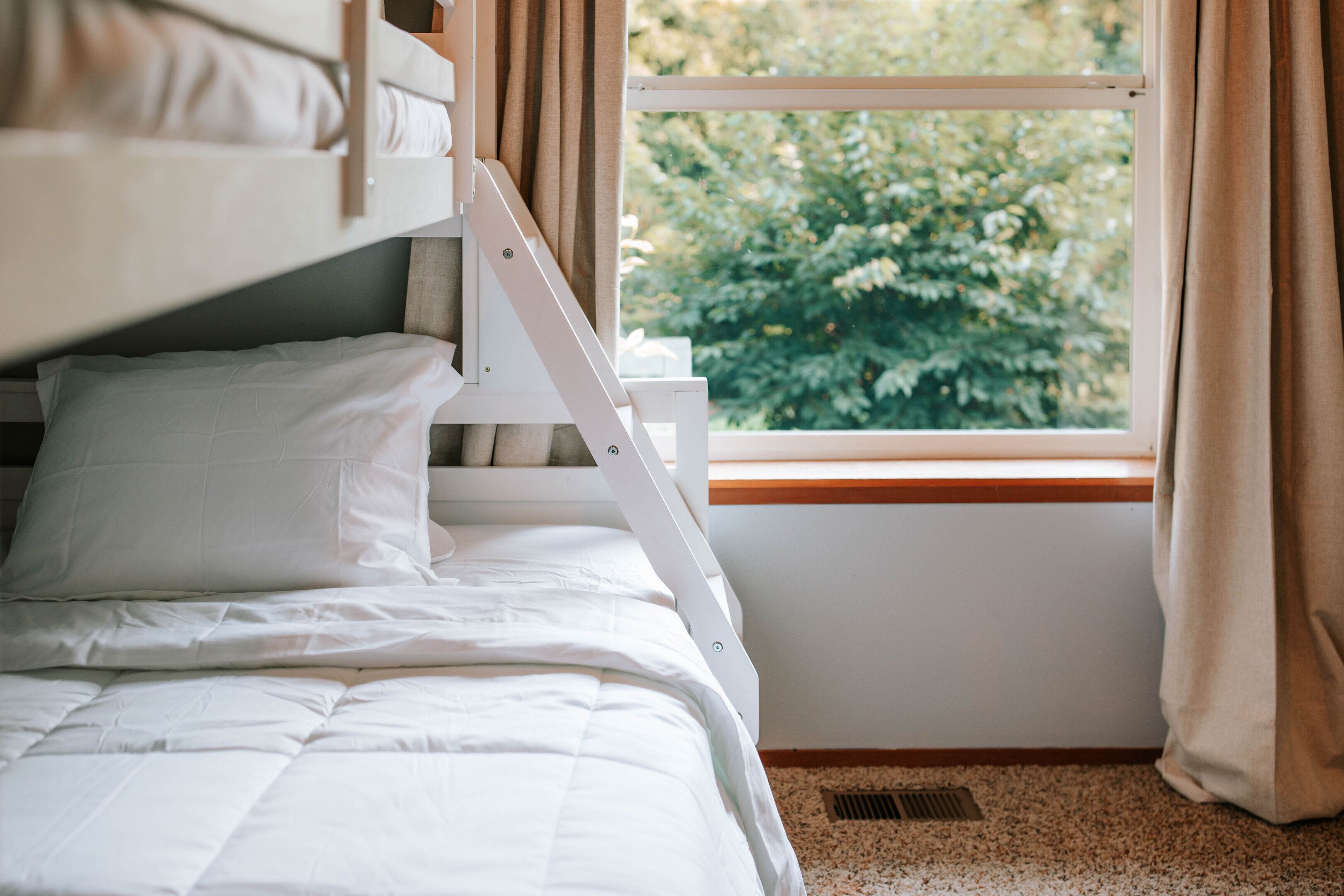 A bunk bed with white bedding, a window with curtains, and a view of greenery outside.