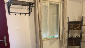 A room with a bench, a window with curtains, a radiator, a shelf with decorative items, and a door. - Apartment in the countryside 30mn from the 24h of Le Mans (Saint-Georges-du-Rosay)