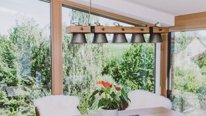 Ein Essbereich mit Holztisch, Topfpflanze und Blick ins Grüne durch große Fenster.
