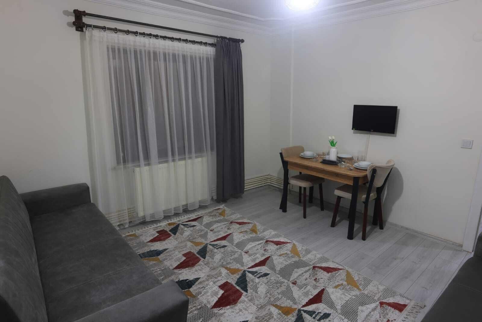 A small, furnished room with a sofa, a dining table with chairs, a TV mounted on the wall, and a patterned rug on the floor.