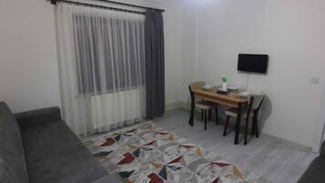 A small, furnished room with a sofa, a dining table with chairs, a TV mounted on the wall, and a patterned rug on the floor.