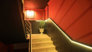 Uma escadaria com uma planta em vaso no topo, corrimão e uma parede com luminária.