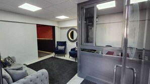 Um espaço de escritório moderno com um sofá cinzento, duas poltronas azuis, um espelho redondo e uma porta de vidro que dá para um banheiro.