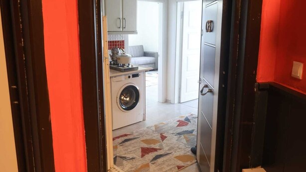 Un couloir menant à une cuisine équipée d’un lave-linge, une porte avec un paillasson portant l’inscription « MAISON », et un mur rouge.